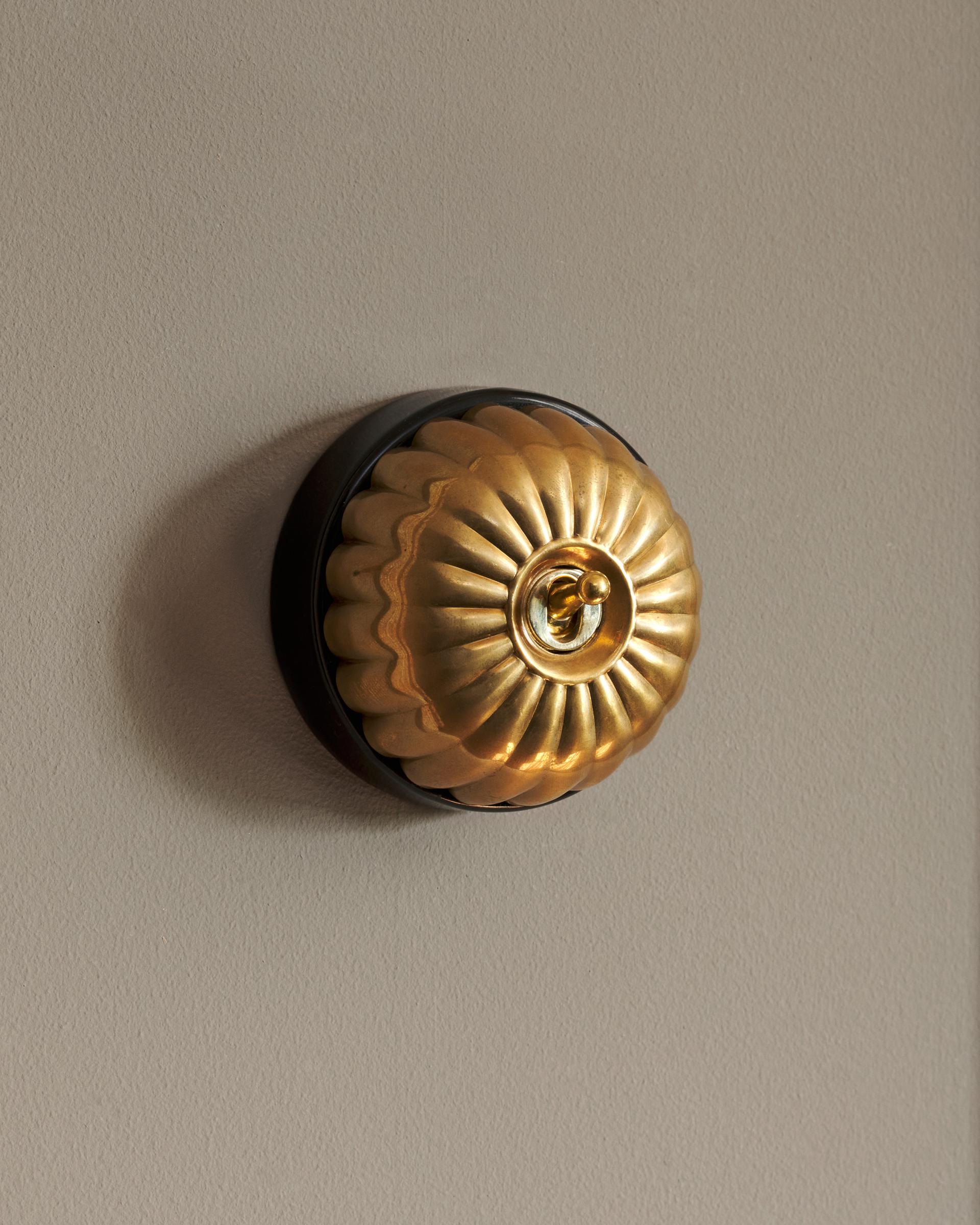 A side-on view of a small 1 gang round-shaped toggle switch in a warm shiny brass finish with a black base.
