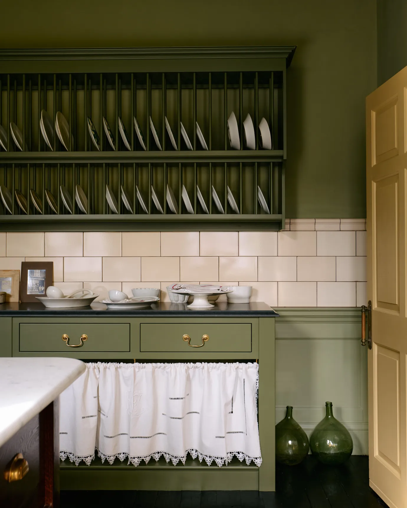 A splashback band of simple cream subway wall tiles runs between a grand open plate rack wall cupboard and a Victorian-style open kitchen cupboard with a slate worktop and ruched linen curtain.