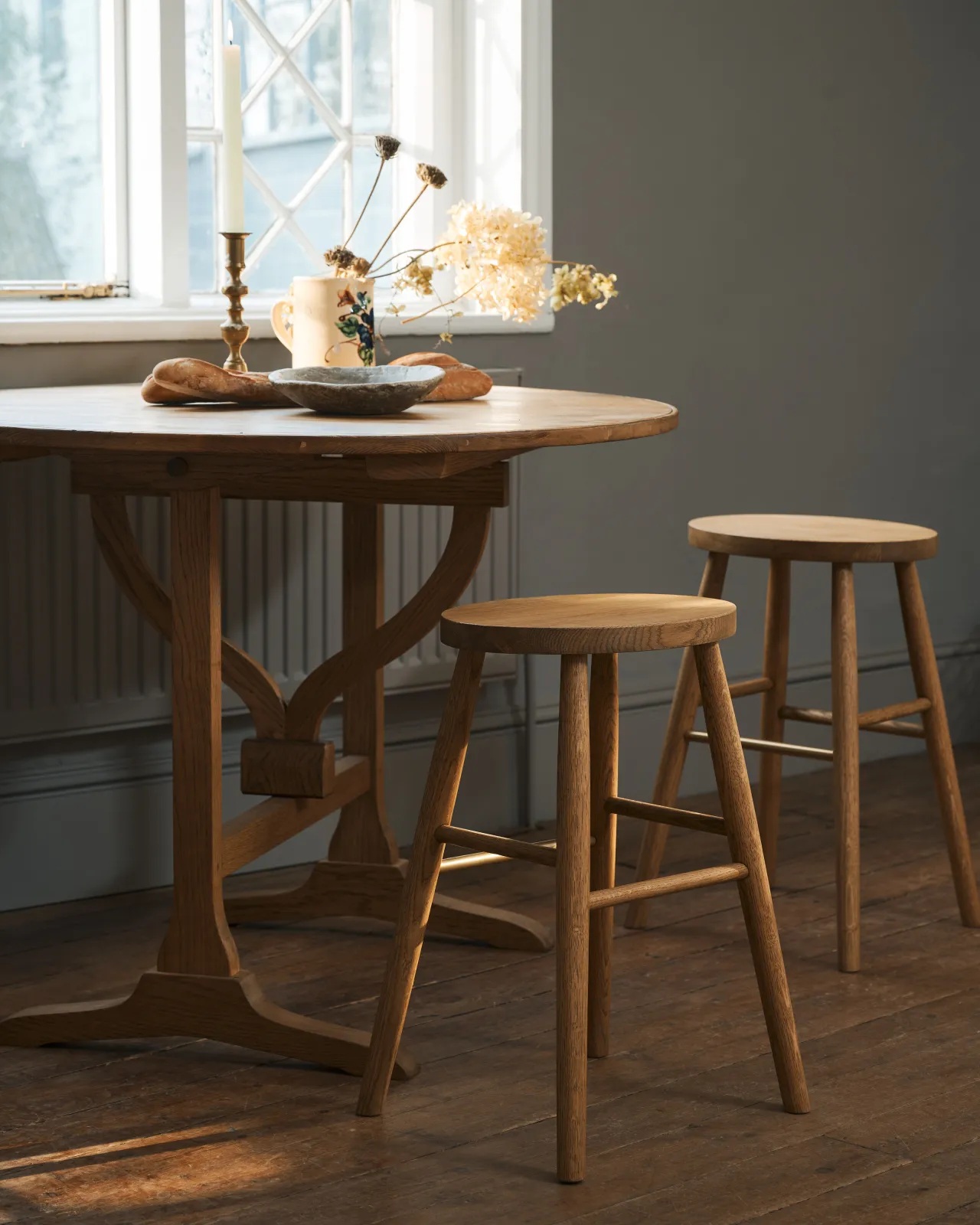 An oblique view of two simple oak milking stools with oval-shaped seats and brass footrails by a round pine table in a dark room.