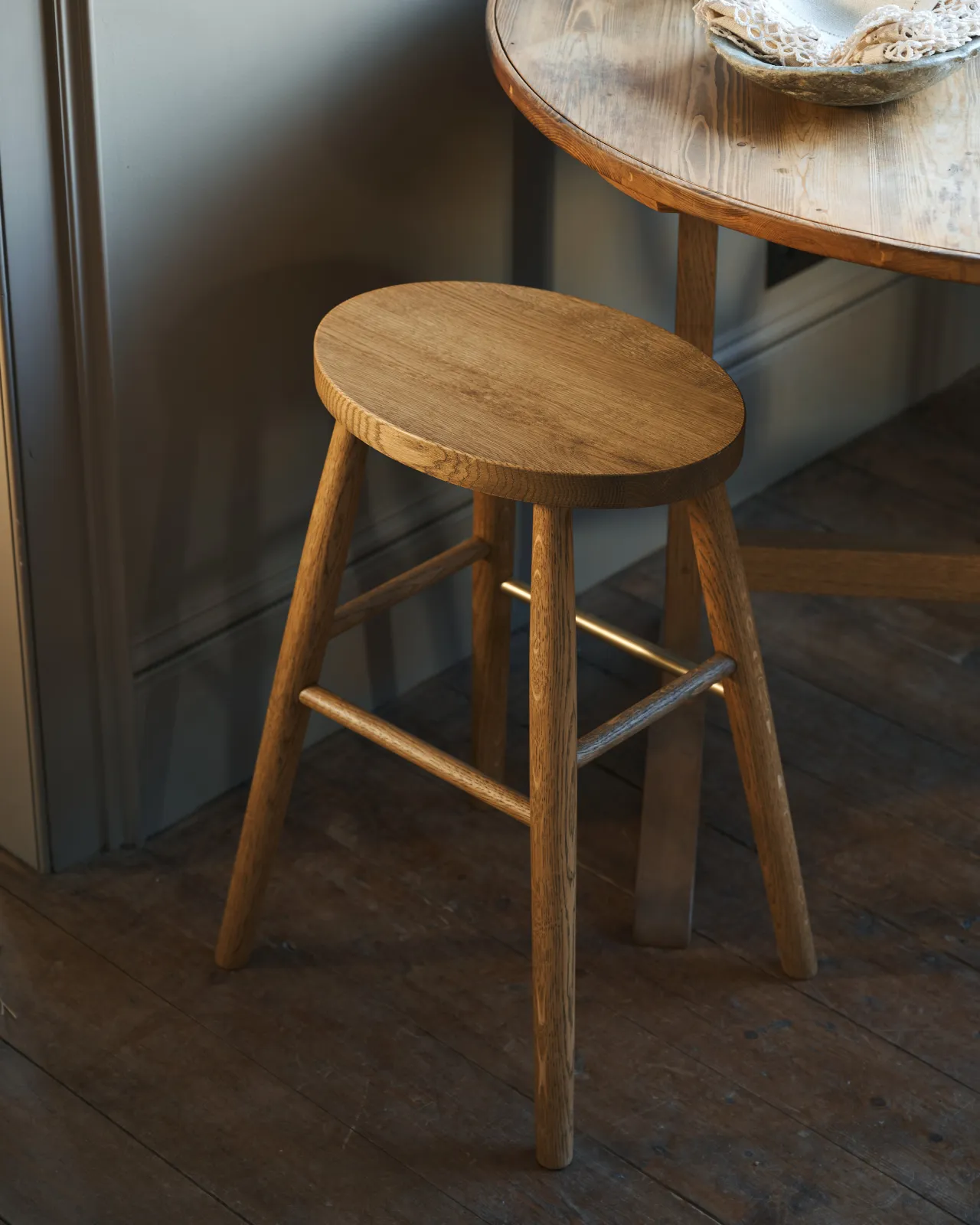 An oblique view of a simple oak milking stool with an oval-shaped seat and a brass footrail.