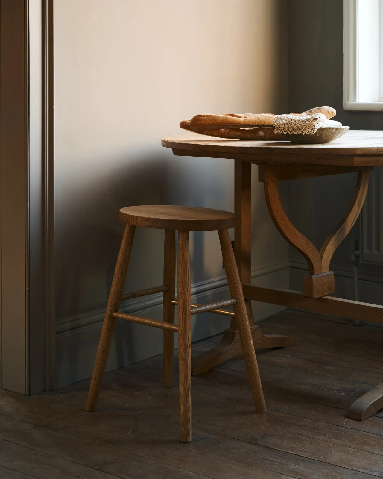 An oblique view of a simple oak milking stool with an oval-shaped seat and a brass footrail by a round pine table in a dark room.