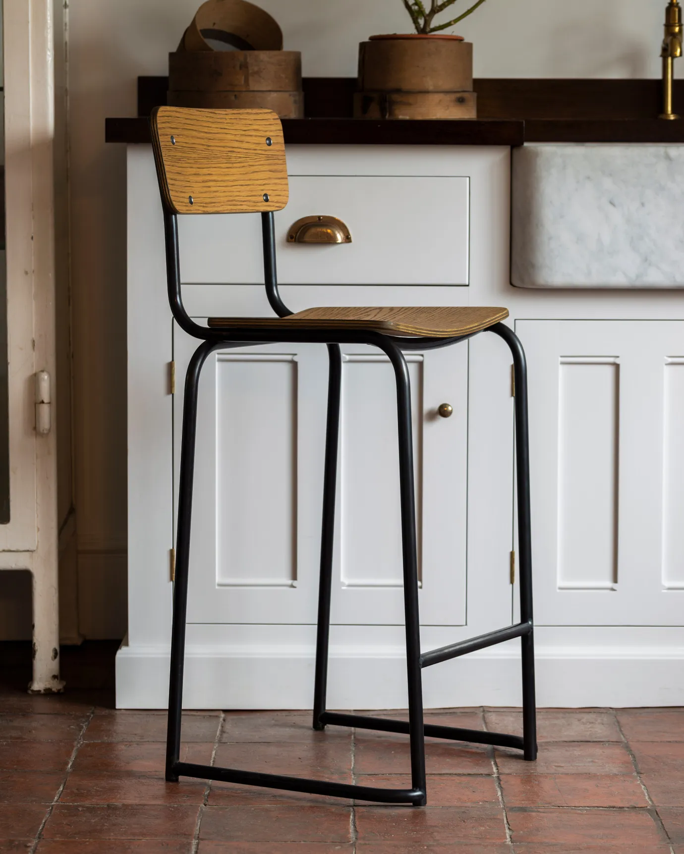 A mid-century-style industrial bar stool with a black tubular metal frame and shaped oak veneer plywood seat and backrest in front of a white painted Georgian-style kitchen sink cupboard.